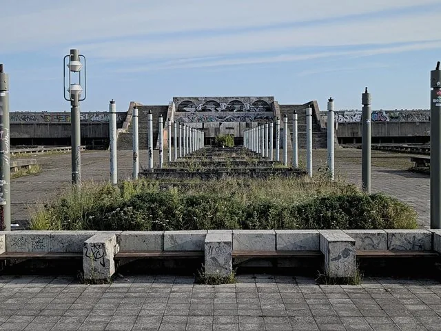 Une esplanade en béton abandonnée et envahie par la végétation avec des lampadaires et des sièges.